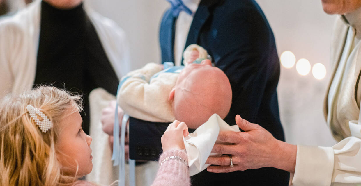 Pieni tyttö kuivaa papin kanssa kastetun vauvan päätä valkoisella liinalla.