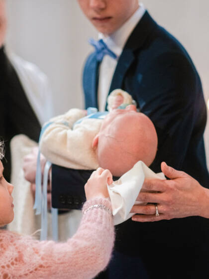 Pieni tyttö kuivaa papin kanssa kastetun vauvan päätä valkoisella liinalla.