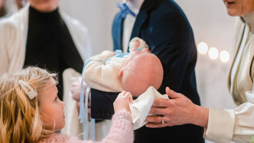 Pieni tyttö kuivaa papin kanssa kastetun vauvan päätä valkoisella liinalla.