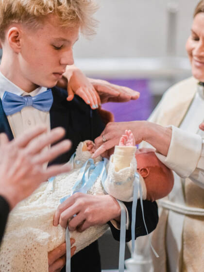 Kastettava vauva on nuoren pojan sylissä, ja pappi ja läheiset ovat nostaneet kätensä siunatakseen vauvaa.