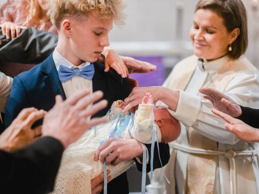 Kastettava vauva on nuoren pojan sylissä, ja pappi ja läheiset ovat nostaneet kätensä siunatakseen vauvaa.