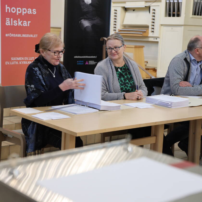 Kolme ihmistä pitkän pöydän ääressä Mikkelin tuomiokirkon etuosassa. Pöydässä vaalikansiot ja kuvan etualalla vaaliuurna.