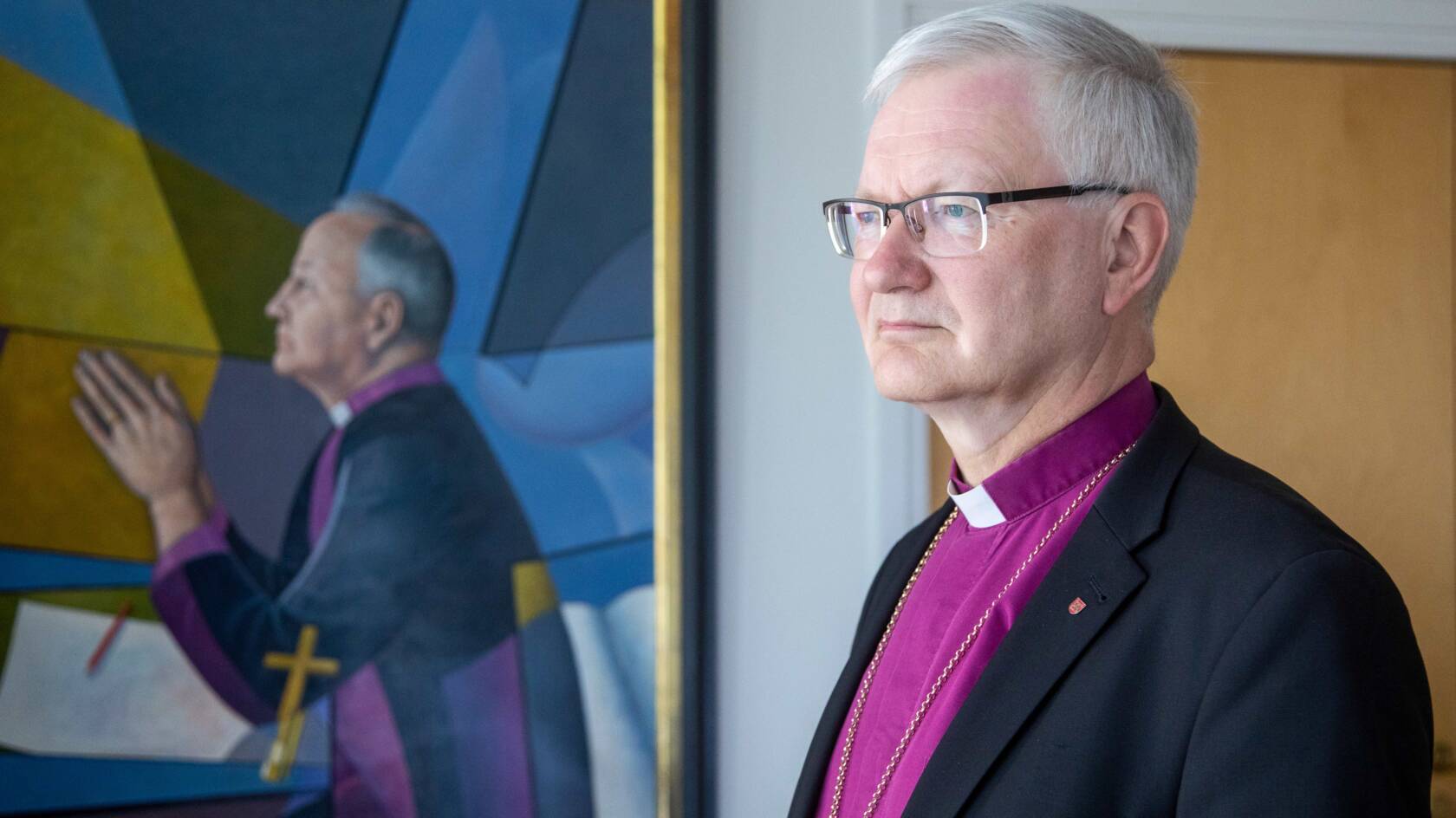 Piispa violetissa pantapaidassaan piispataulun vieressä
