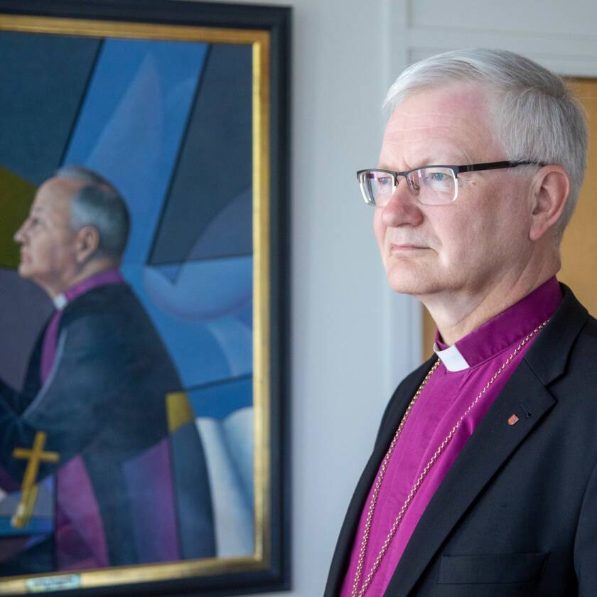 Piispa violetissa pantapaidassaan piispataulun vieressä