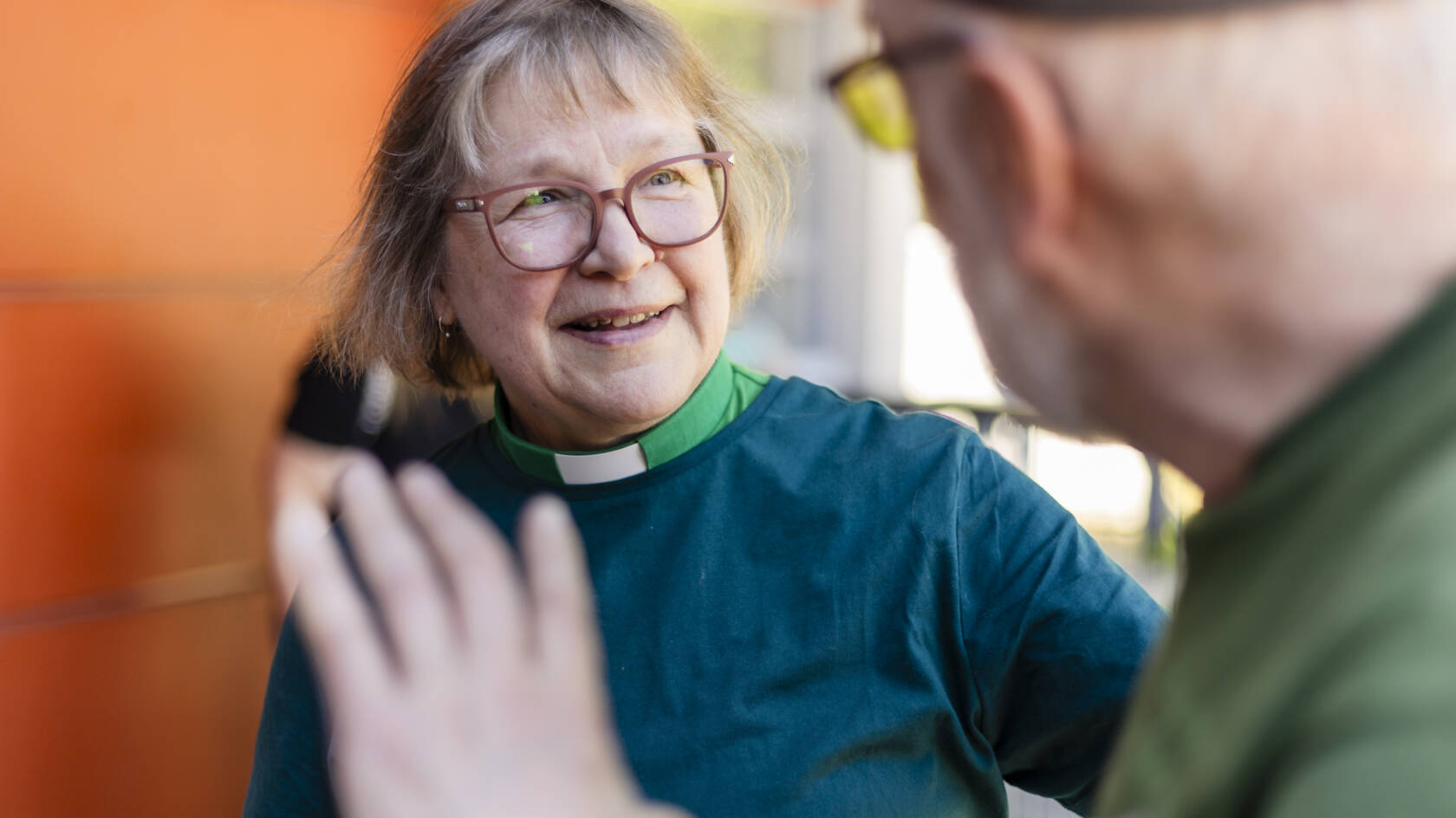 Nainen, jolla on silmälasit ja vihreä paita, hymyilee ja keskustelee toisen henkilön kanssa ulkona. Taustalla näkyy oranssi seinä.
