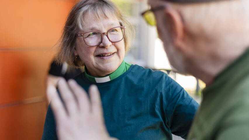 Nainen, jolla on silmälasit ja vihreä paita, hymyilee ja keskustelee toisen henkilön kanssa ulkona. Taustalla näkyy oranssi seinä.