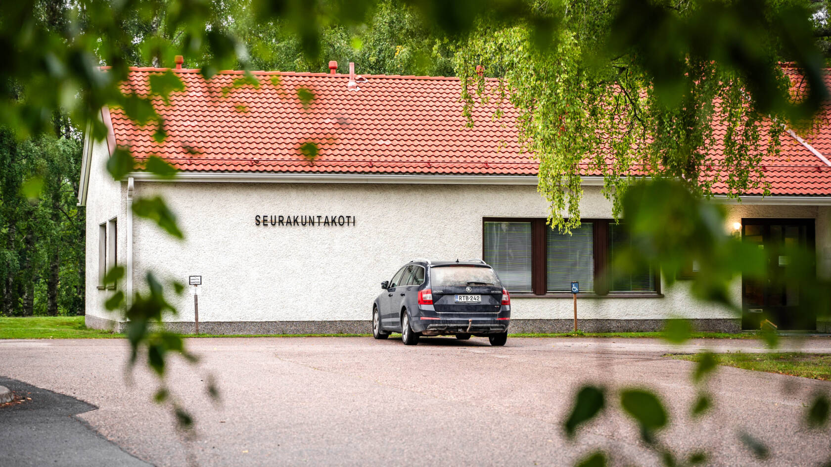 Valkorapattu punatiilikattoinen rakennus, jonka seinässä lukee seurakuntakoti. Rakennuksen pihassa auto. Kuvan etualalla puun oksia.